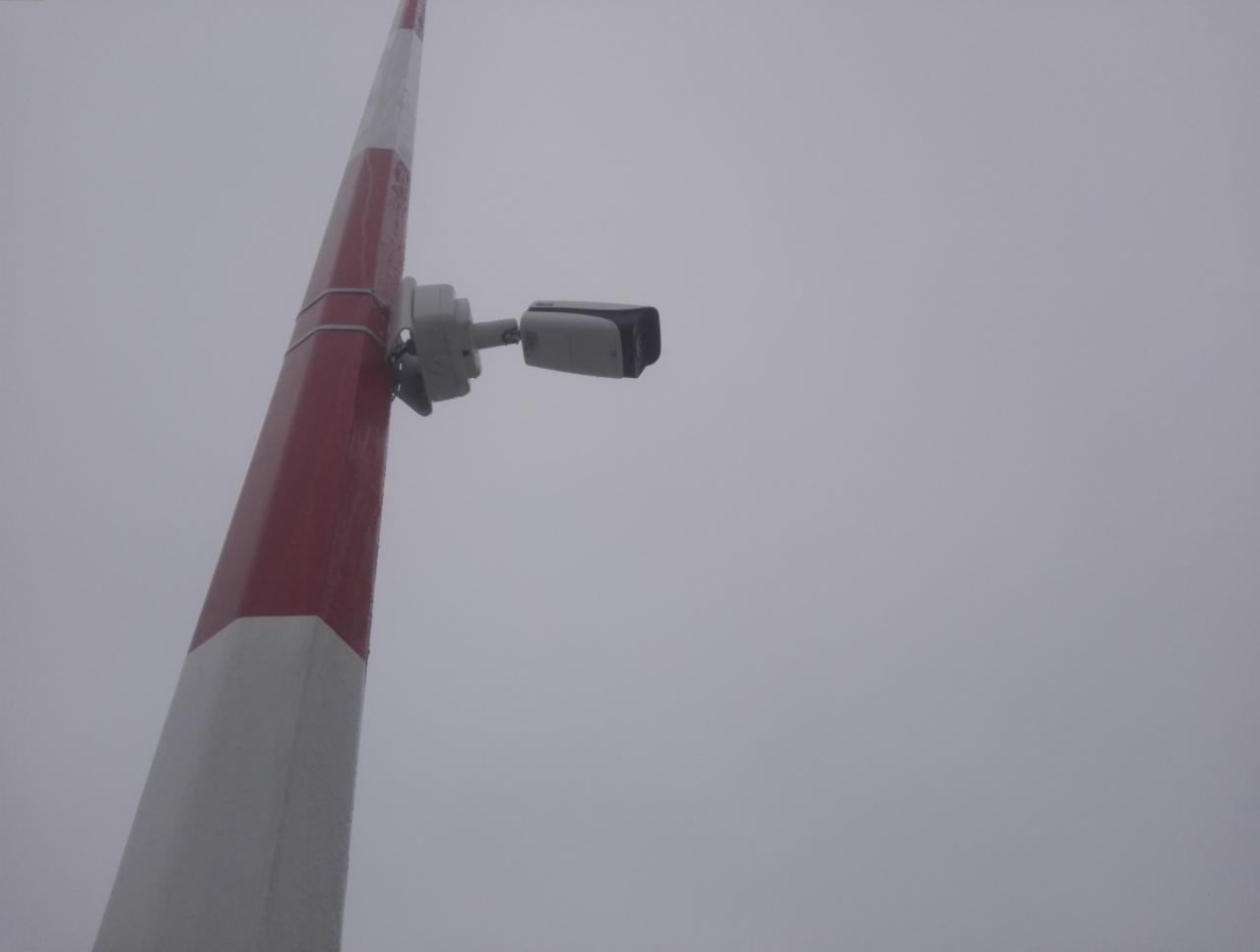 Kamera monitoringu przymocowana do słupka szlabanu pomalowanego w biało-czerwone pasy, widok z dołu na tle pochmurnego nieba.