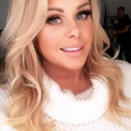 Portret uśmiechniętej blondynki w białym swetrze na tle studia fotograficznego z widocznym oświetleniem i osobą obsługującą sprzęt.