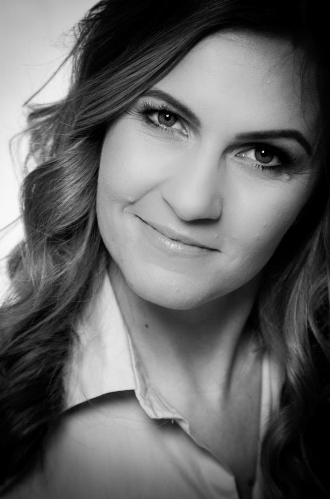 Portret uśmiechniętej kobiety z długimi, kręconymi włosami, w czarno-białej tonacji, z widocznymi zmarszczkami mimicznymi wokół oczu.