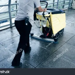 Pracownik w czarnych spodniach obsługuje żółtą maszynę czyszczącą posadzkę w przestrzeni publicznej, widoczne mokre płytki i metalowe barierki.
