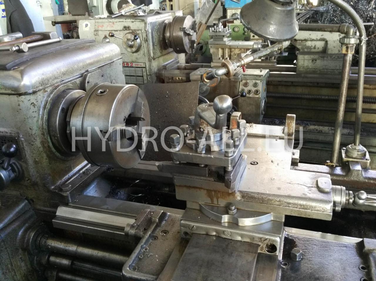 Stara tokarka z uchwytem szczękowym i zamocowanym narzędziem skrawającym, oświetlona lampą warsztatową, w tle widoczna kolejna tokarka.