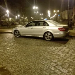 Srebrny samochód taxi marki Mercedes zaparkowany nocą na mokrym, brukowanym placu, oświetlonym przez latarnie.