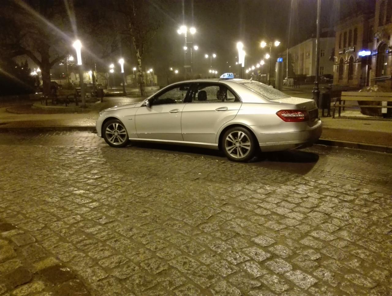 Srebrny samochód taxi marki Mercedes zaparkowany nocą na mokrym, brukowanym placu, oświetlonym przez latarnie.