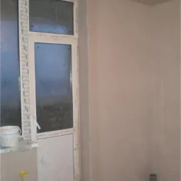 Świeżo otynkowane wnętrze z oknem balkonowym, wiaderkiem z zaprawą na parapecie i odpływem kanalizacyjnym w rogu.