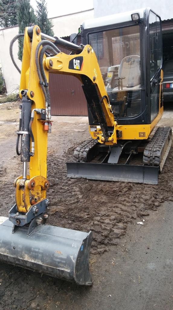 Żółta minikoparka CAT z łyżką na błotnistym terenie, widoczny budynek w tle.