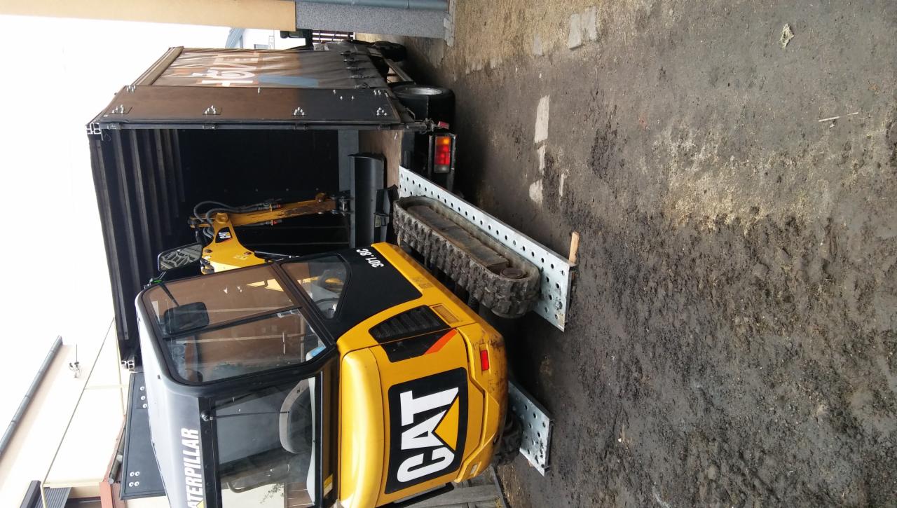 Żółta mini koparka CAT 301.8C wjeżdża po rampie do wnętrza ciężarówki z plandeką; widoczne gąsienice i kabina maszyny.