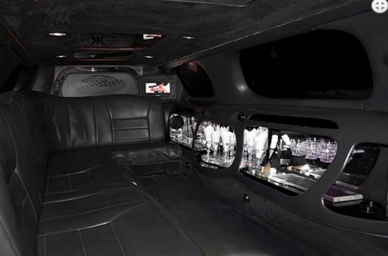 Wnętrze luksusowej limuzyny z czarną skórzaną tapicerką, barkiem z podświetlanymi szklankami i butelkami, oraz wbudowanym ekranem telewizyjnym.