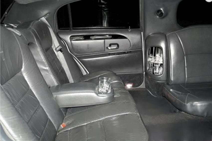 Wnętrze limuzyny z czarną skórzaną tapicerką, szklanka z napojem w podłokietniku, widok na tylne siedzenia i drzwi.