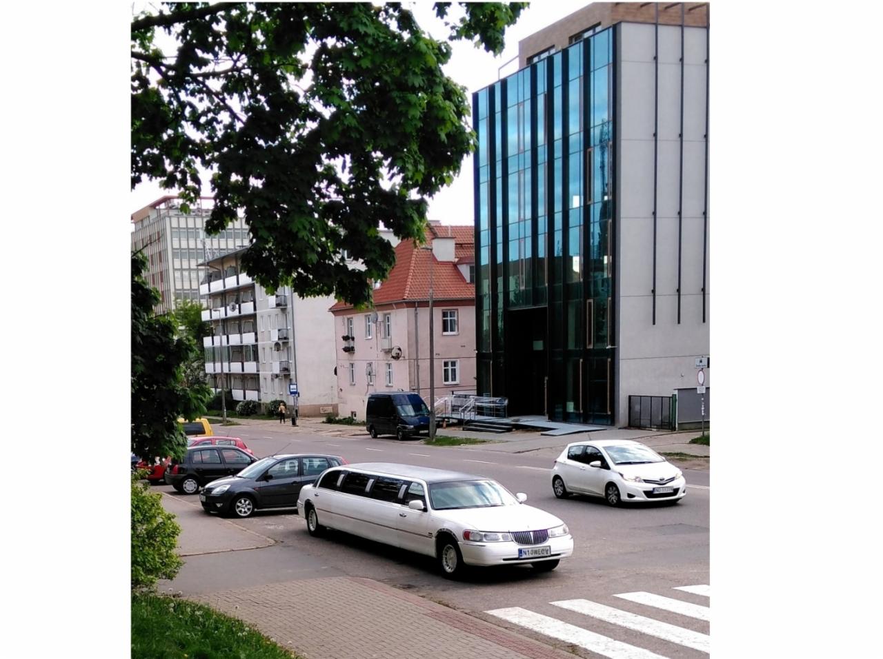 Biała limuzyna Lincoln na skrzyżowaniu w mieście, w tle budynki biurowe i mieszkalne.