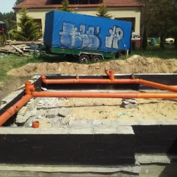 Fundamenty domu w trakcie budowy z widoczną hydroizolacją i systemem rur kanalizacyjnych w kolorze pomarańczowym, na zewnątrz widoczna przyczepa z plandeką z graffiti.
