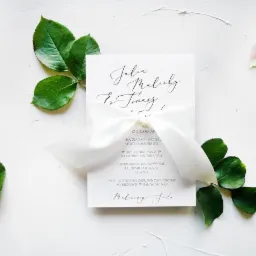 Eleganckie zaproszenie ślubne z kaligrafowanym imieniem i nazwiskiem, przewiązane białą wstążką, otoczone zielonymi liśćmi i płatkiem róży na białym tle.