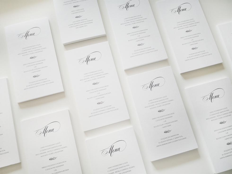 Eleganckie, minimalistyczne karty menu z czarnym, kaligraficznym napisem 'Menu' i listą dań na białym tle, ułożone pod kątem na jasnej powierzchni.