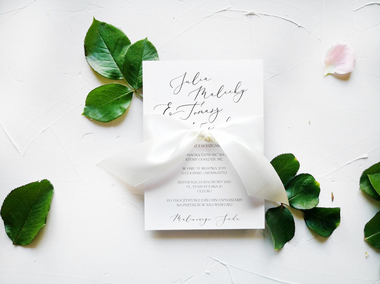 Eleganckie zaproszenie ślubne z kaligrafowanym imieniem i nazwiskiem, przewiązane białą wstążką, otoczone zielonymi liśćmi i płatkiem róży na białym tle.