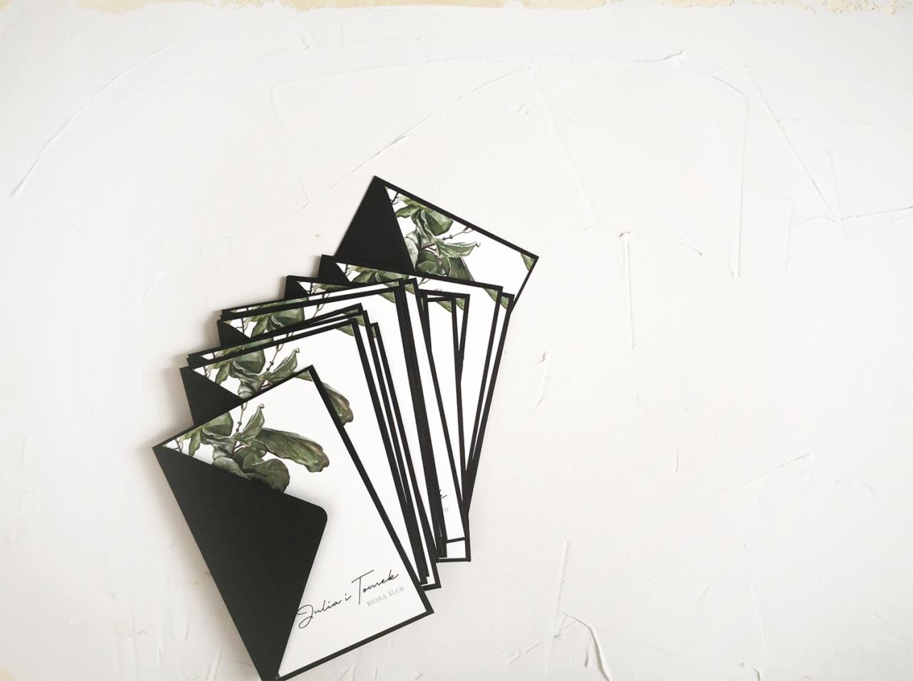 Stos eleganckich zaproszeń ślubnych z motywem zielonych liści i czarnymi kopertami, ułożonych na białym, teksturowanym tle.