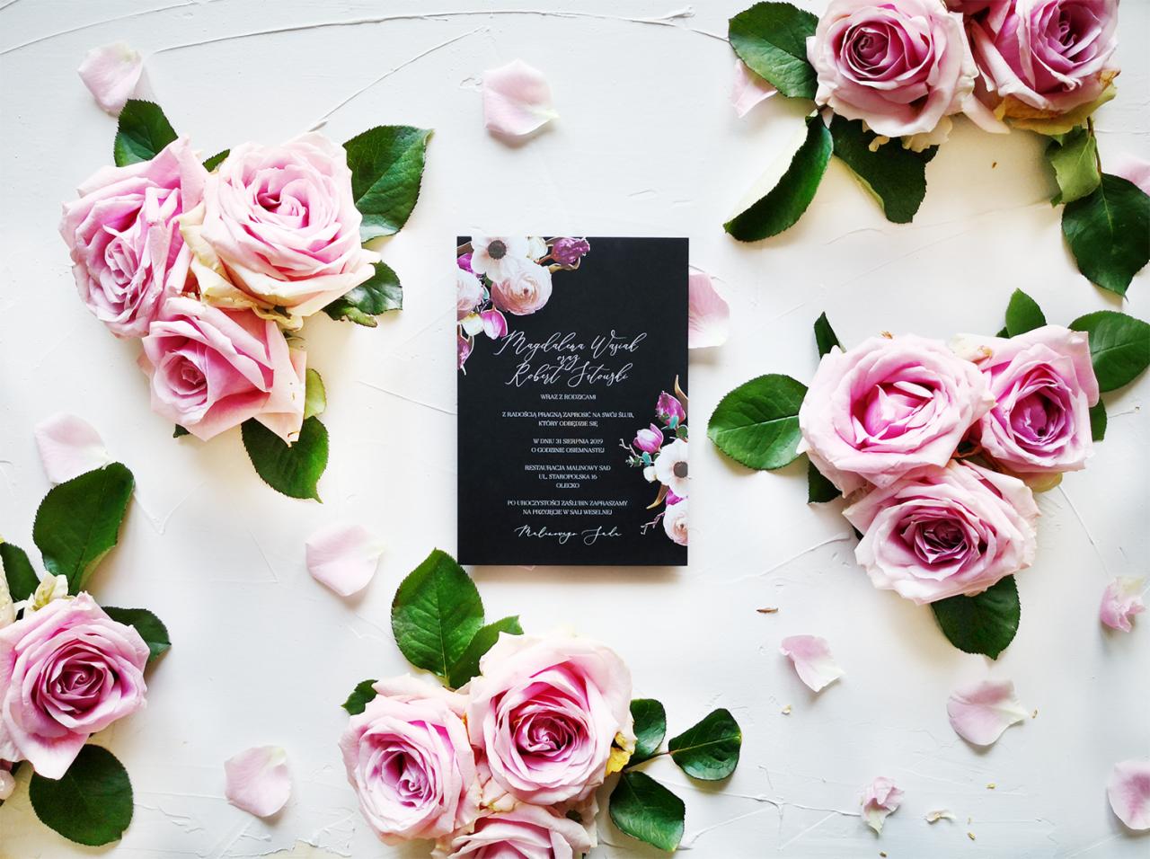 Eleganckie, czarne zaproszenie ślubne z kaligrafią i motywem kwiatowym, otoczone różowymi różami i płatkami na białym tle.