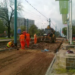 Prace ziemne przy torach tramwajowych w mieście. Robotnicy w odblaskowych strojach oraz koparka w trakcie prac. W tle budynki mieszkalne i słupy trakcyjne.