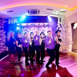 Zespół w fioletowych koszulach i czarnych strojach pozuje z uniesionymi kciukami na scenie z dekoracjami balonowymi i napisem 'HK' w tle.