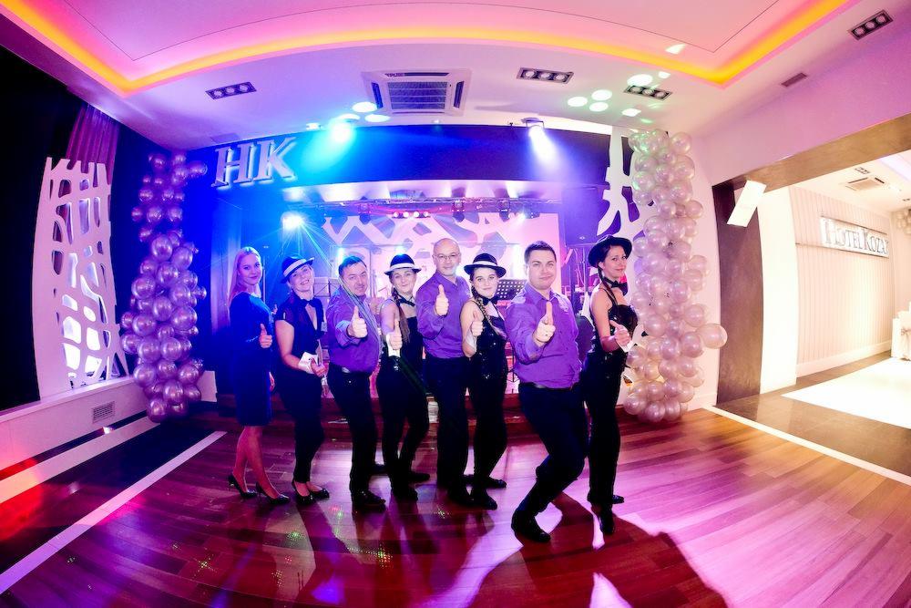 Zespół w fioletowych koszulach i czarnych strojach pozuje z uniesionymi kciukami na scenie z dekoracjami balonowymi i napisem 'HK' w tle.
