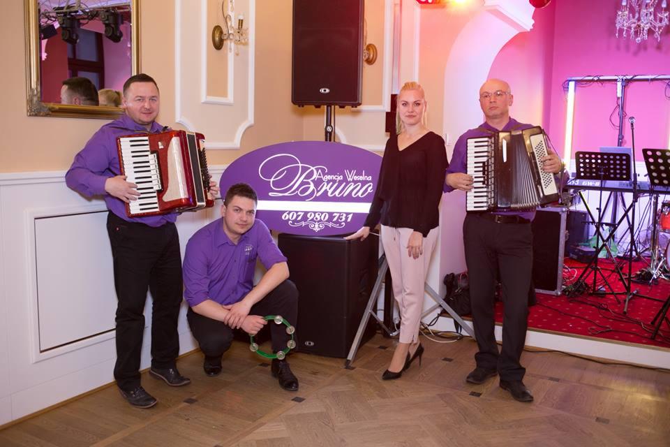 Zespół muzyczny w fioletowych koszulach z akordeonami i tamburynem prezentuje się na tle sceny z logo Agencji Weselnej Bruno.