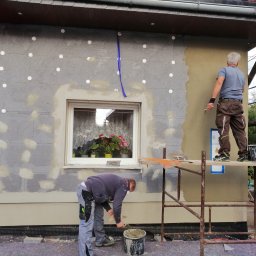 MarekSzałacki - Ocieplanie elewacji domu jednorodzinnego. Dwóch mężczyzn na rusztowaniu nakłada tynk. Widoczne białe kołki styropianowe i okno z kwiatami.