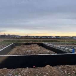 MarekSzałacki - Zabezpieczone fundamenty domu z bloczków betonowych, pokryte czarną izolacją, na tle pola i zabudowań w oddali, pod pochmurnym niebem o zmierzchu.