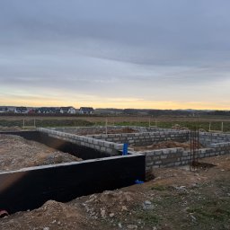 MarekSzałacki - Fundamenty domu w trakcie budowy: ściany z bloczków betonowych, izolacja przeciwwilgociowa, zbrojenie. W tle krajobraz wiejski i zachodzące słońce.