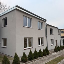 MarekSzałacki - Dwukondygnacyjny budynek po remoncie elewacji w odcieniach szarości, z białymi oknami i rynną. Wzdłuż ścieżki posadzone małe drzewka iglaste.