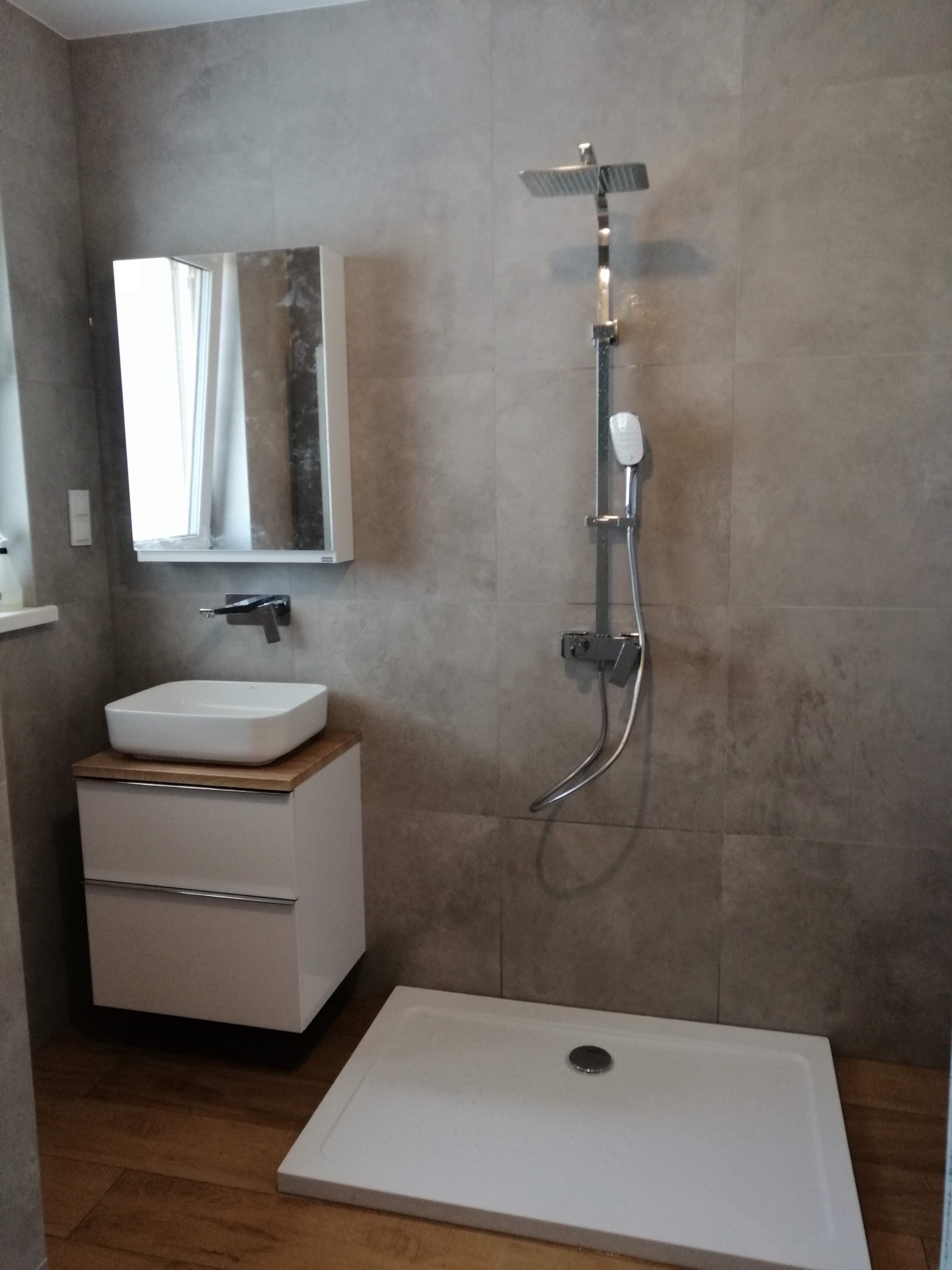 Nowoczesna łazienka po remoncie: szare płytki, biała umywalka z szafką z drewnianym blatem i kabina prysznicowa. Minimalistyczny design, widok na całe pomieszczenie.