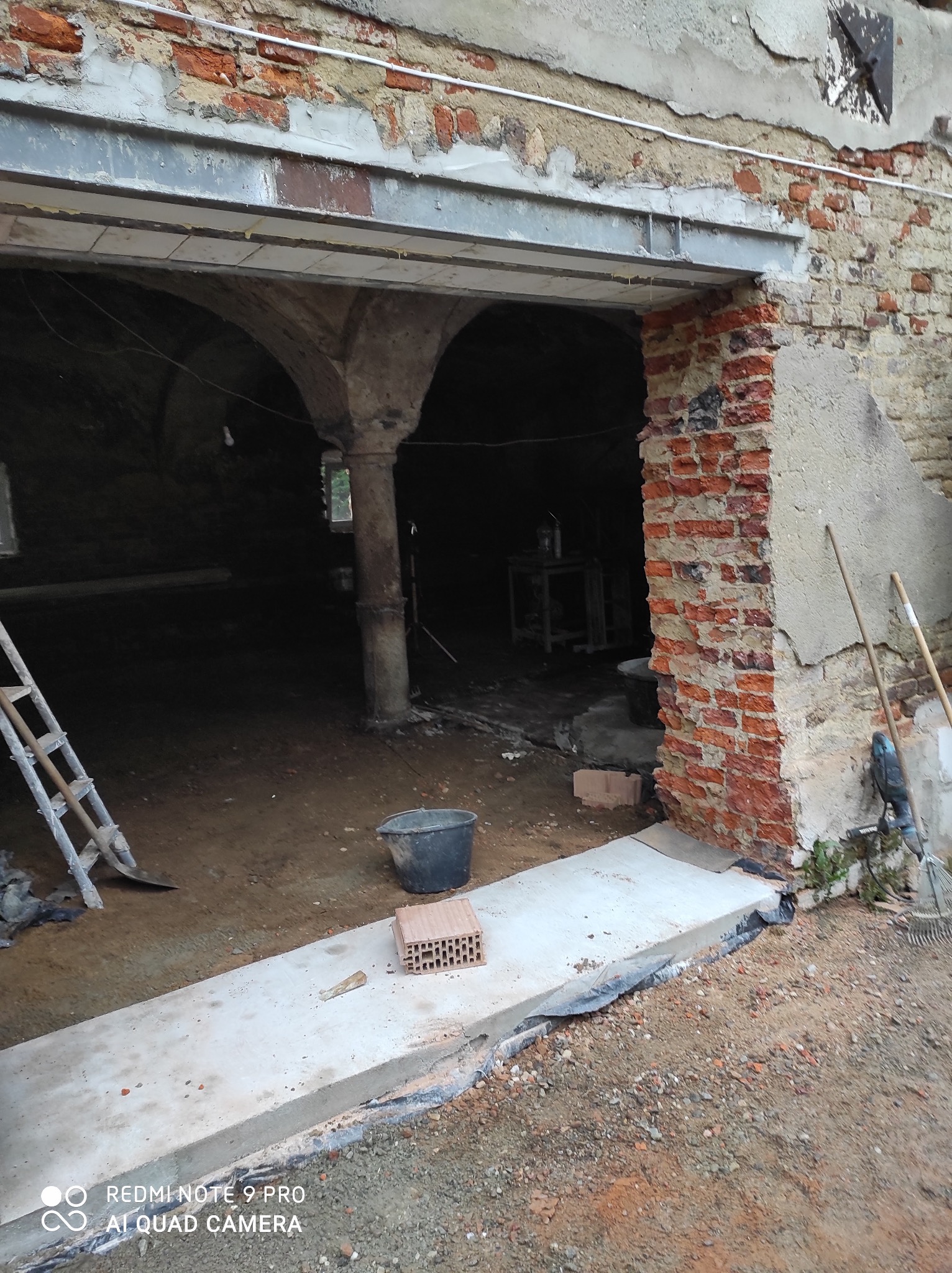 Wnętrze kamienicy w trakcie remontu: odsłonięta cegła, kolumny, widoczne narzędzia i materiały budowlane. Surowy stan przed wykończeniem.