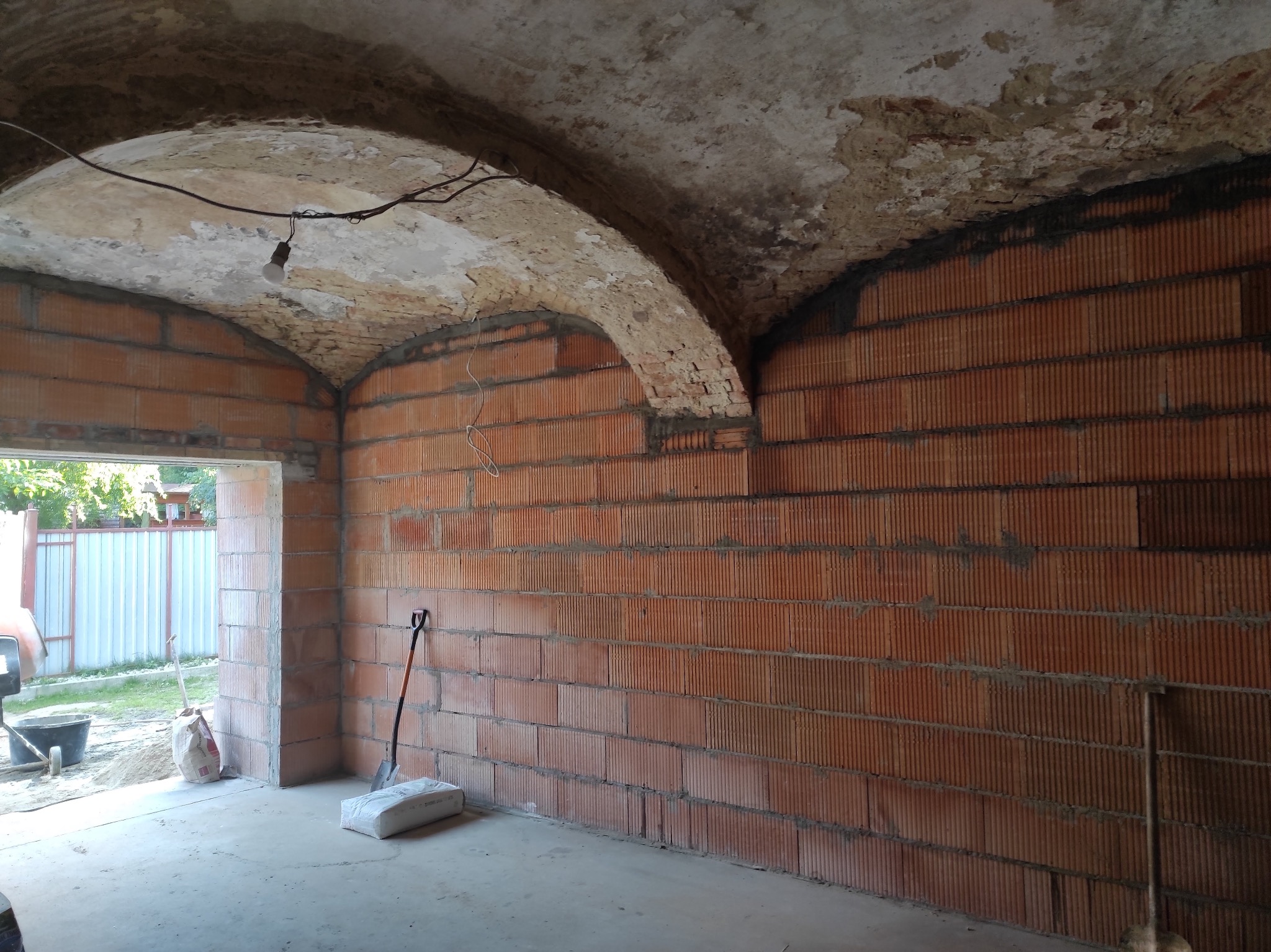 Wnętrze w trakcie budowy: ściany z czerwonej cegły, sklepienie łukowe z odsłoniętą cegłą, worki z zaprawą i narzędzia budowlane. Widok na zewnątrz przez otwarte drzwi.