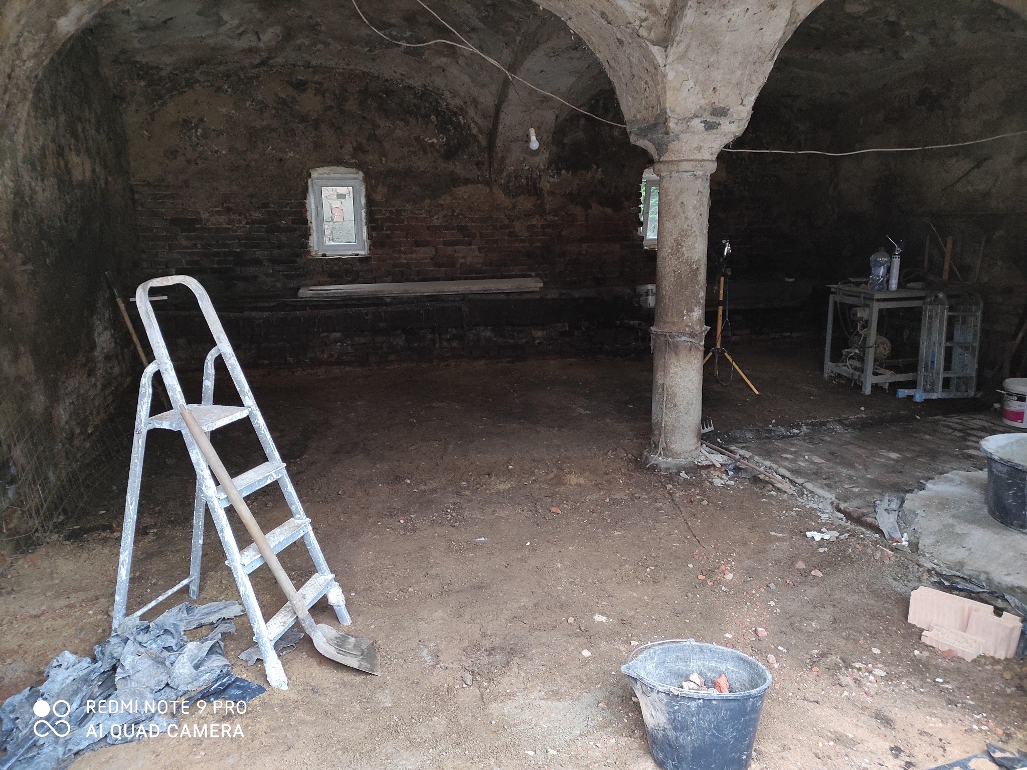 Piwnica w trakcie remontu: ceglane ściany, sklepienie łukowe, drabina, narzędzia i wiadro. Przygotowanie do malowania lub wykończenia wnętrza.