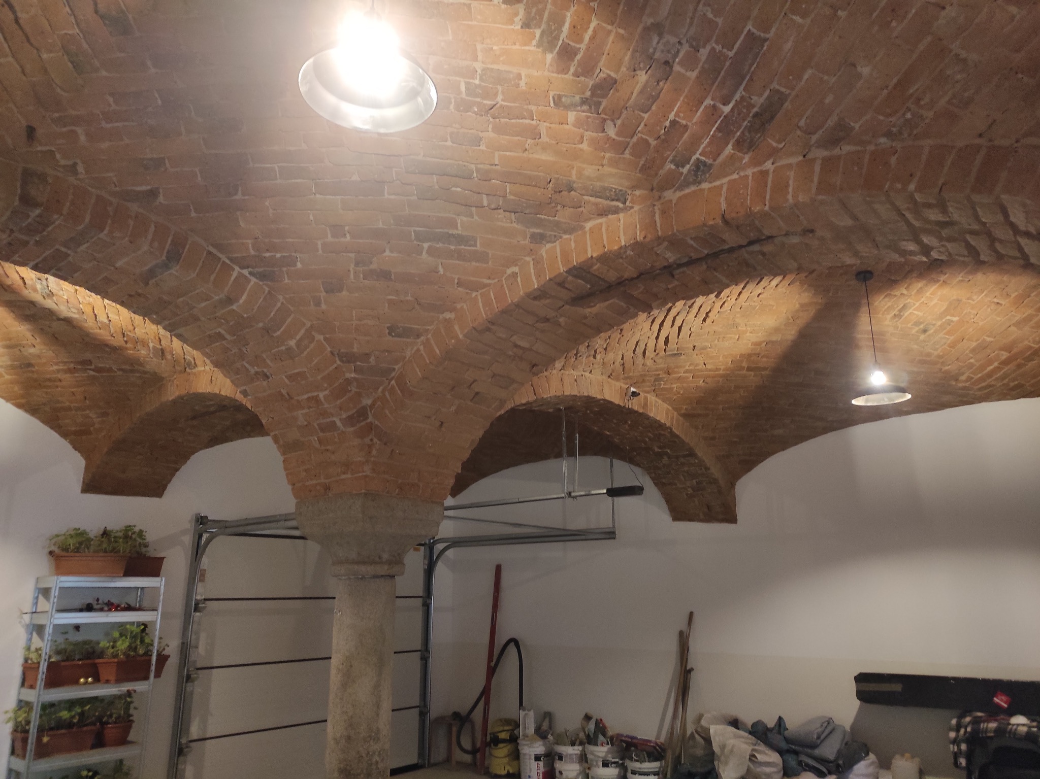 Ceglany, łukowy sufit z oświetleniem w pomieszczeniu z kolumną i regałem z roślinami. Widoczne narzędzia i puszki farb. Charakterystyczna architektura wnętrza.