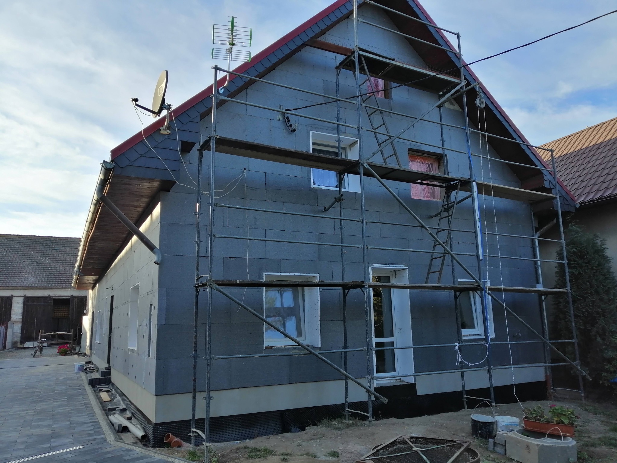 Dom w trakcie ocieplania styropianem, widoczne rusztowania, okna zabezpieczone folią. Część elewacji już ocieplona, część w trakcie prac. Dach pokryty szarą dachówką.