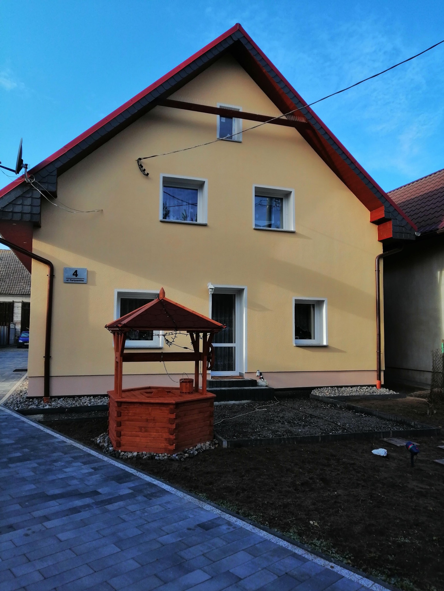 Odnowiona elewacja domu z beżową farbą, czerwonym dachem i studnią z drewna. Widoczne okna z białymi ramami i świeżo przygotowany teren pod trawnik.