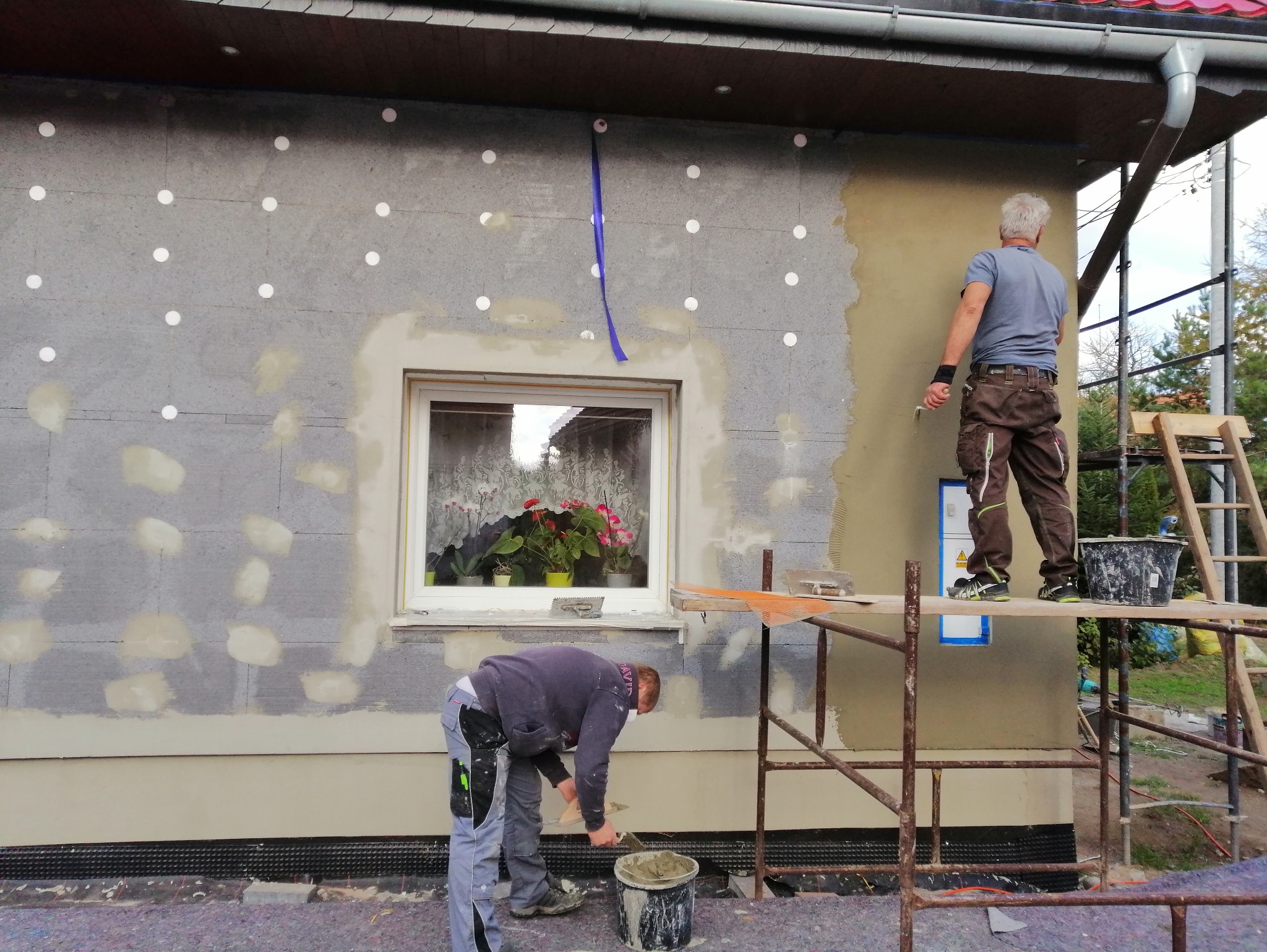 Ocieplanie elewacji domu jednorodzinnego. Dwóch mężczyzn na rusztowaniu nakłada tynk. Widoczne białe kołki styropianowe i okno z kwiatami.