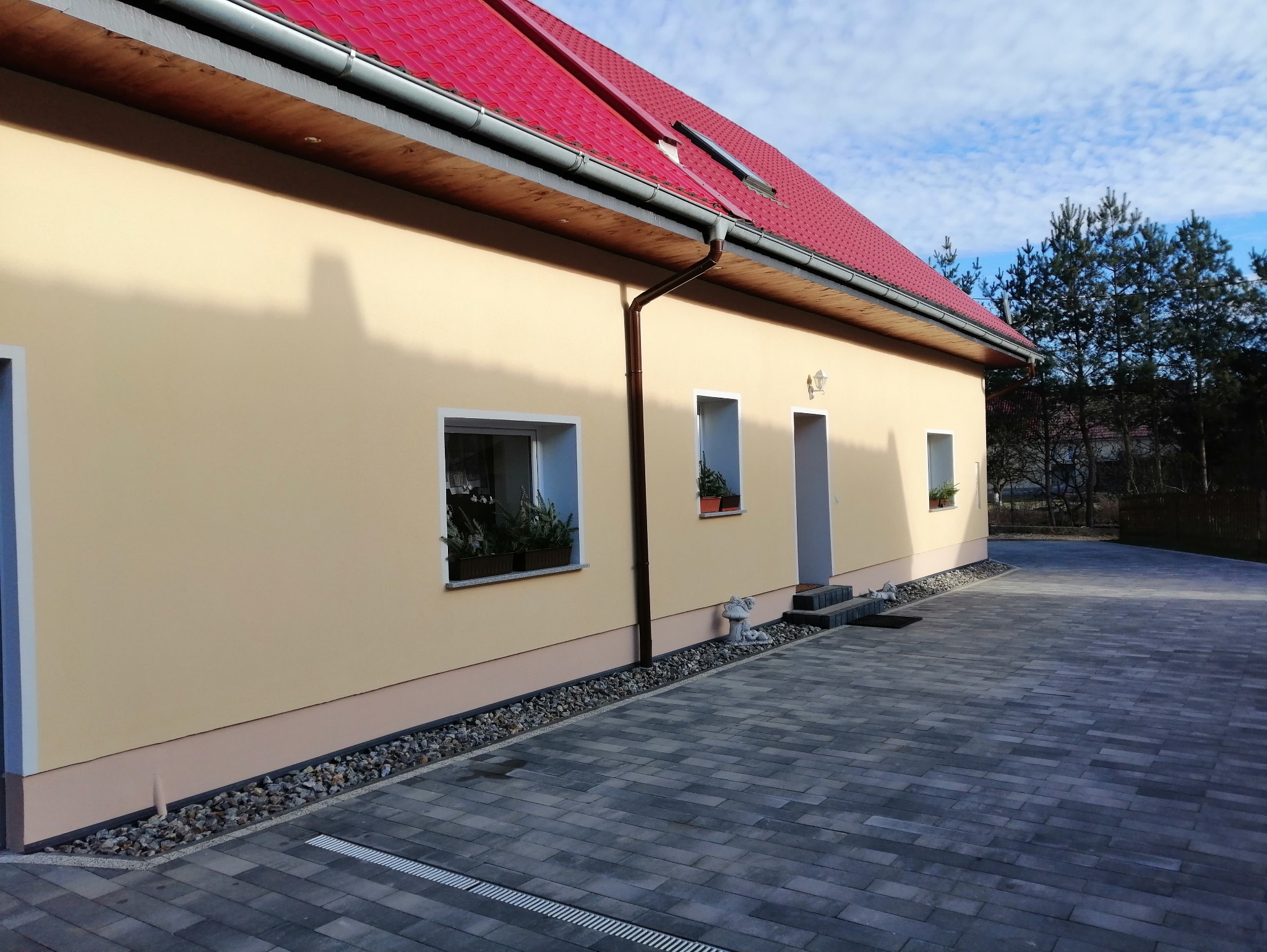 Odnowiona elewacja domu z czerwonym dachem, widok z boku. Jasna fasada, okna z kwiatami, kostka brukowa i ozdobne kamienie przy ścianie. Słoneczny dzień.