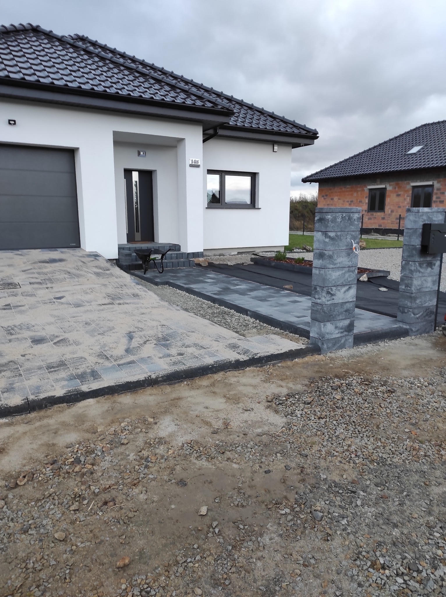 Ułożona kostka brukowa na podjeździe i chodniku w Prusinowicach. Widoczne słupki z kostki i fragment domu z garażem. Prace brukarskie w toku.