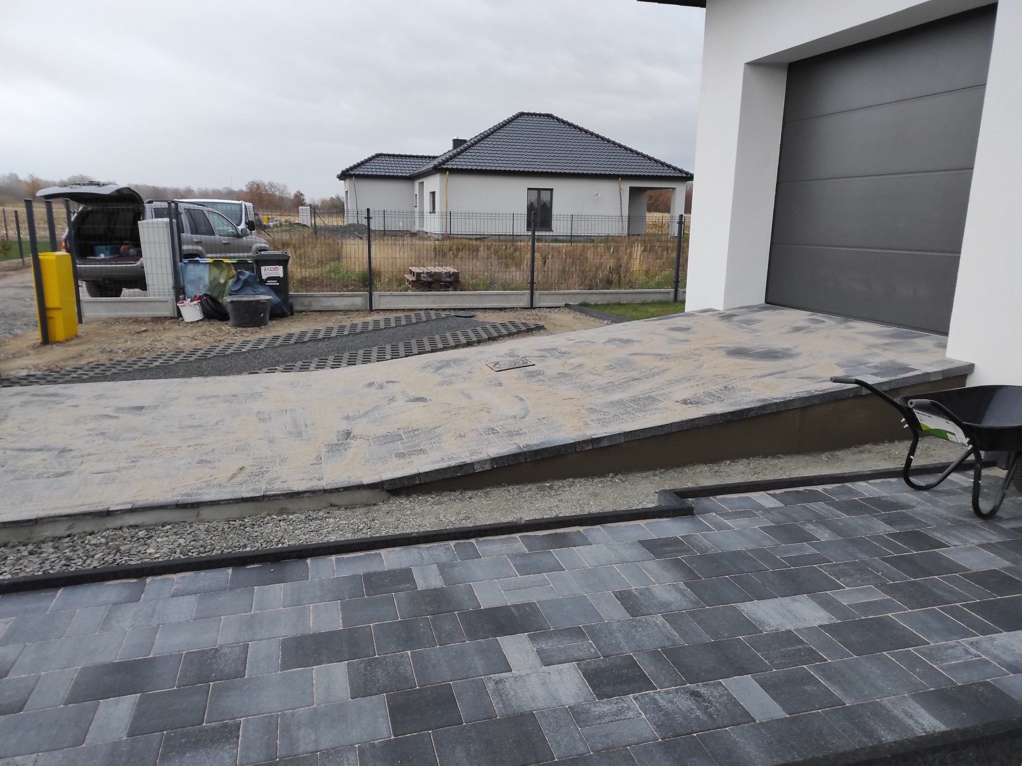Układanie kostki brukowej w odcieniach szarości na podjeździe do garażu. Widoczny fragment domu z szarą bramą garażową i trawnik w tle.