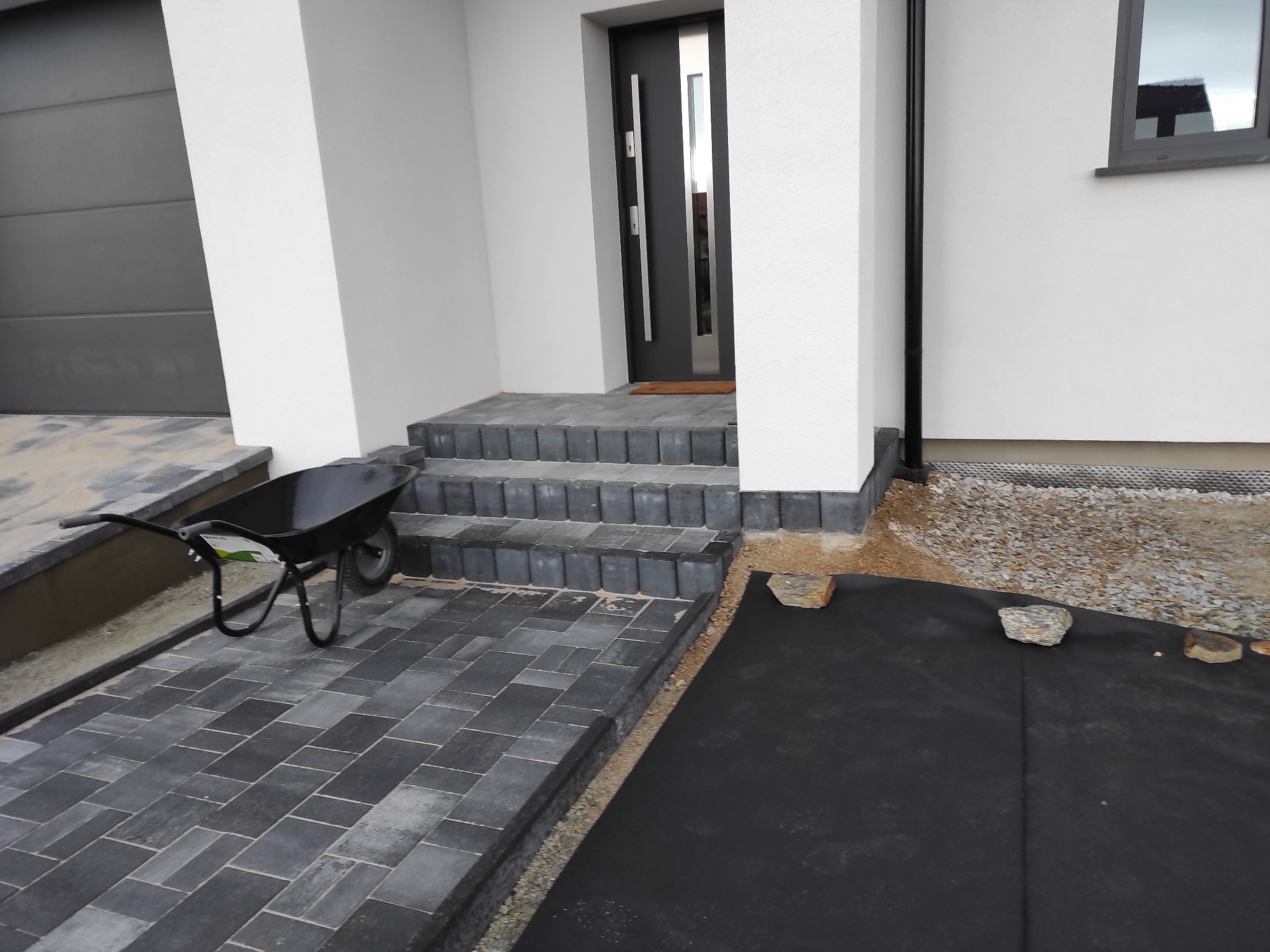 Ułożona kostka brukowa przed wejściem do domu, widoczna taczka i fragment folii. Nowoczesne wykończenie z szarej kostki.