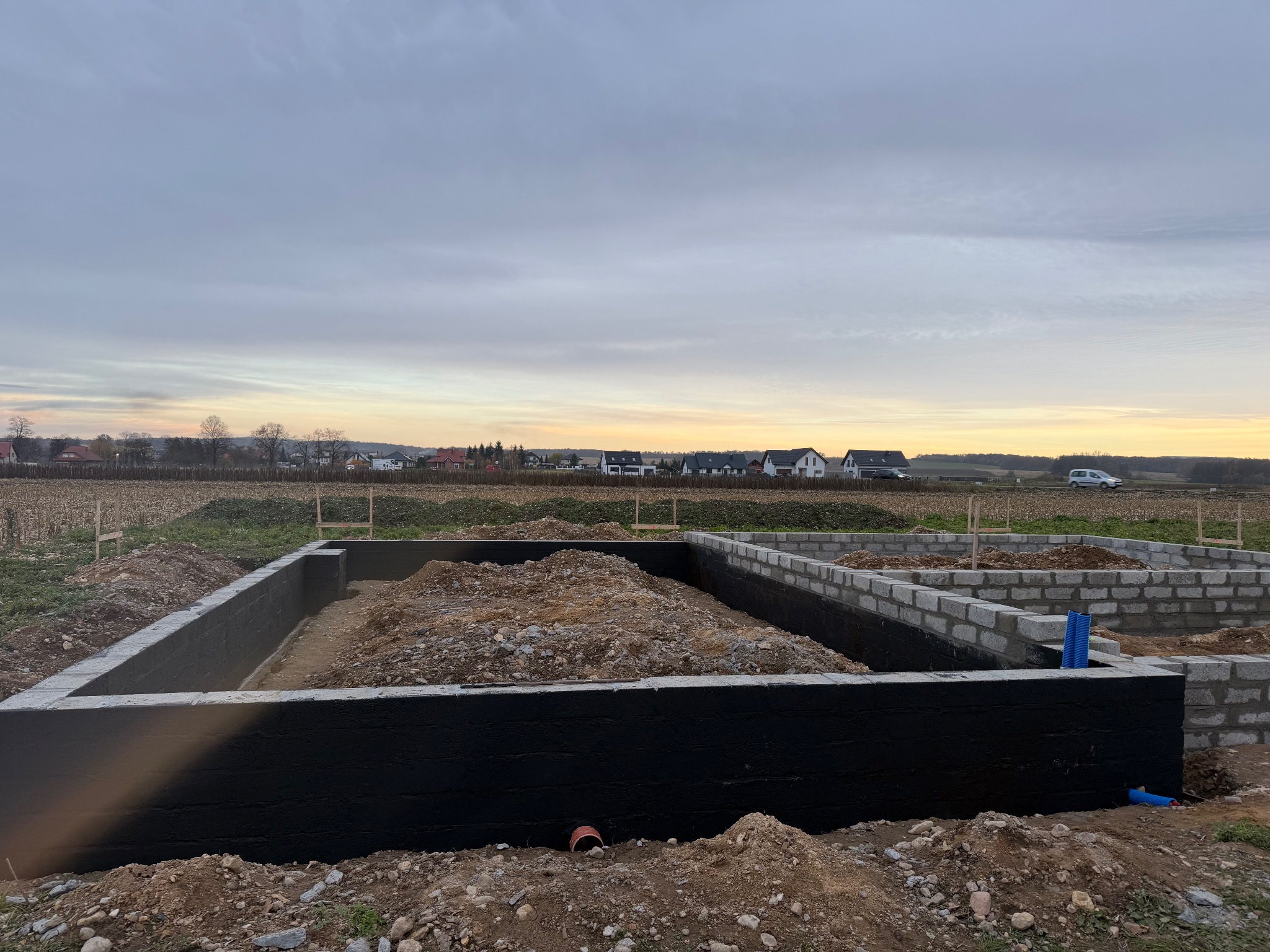 Zabezpieczone fundamenty domu z bloczków betonowych, pokryte czarną izolacją, na tle pola i zabudowań w oddali, pod pochmurnym niebem o zmierzchu.