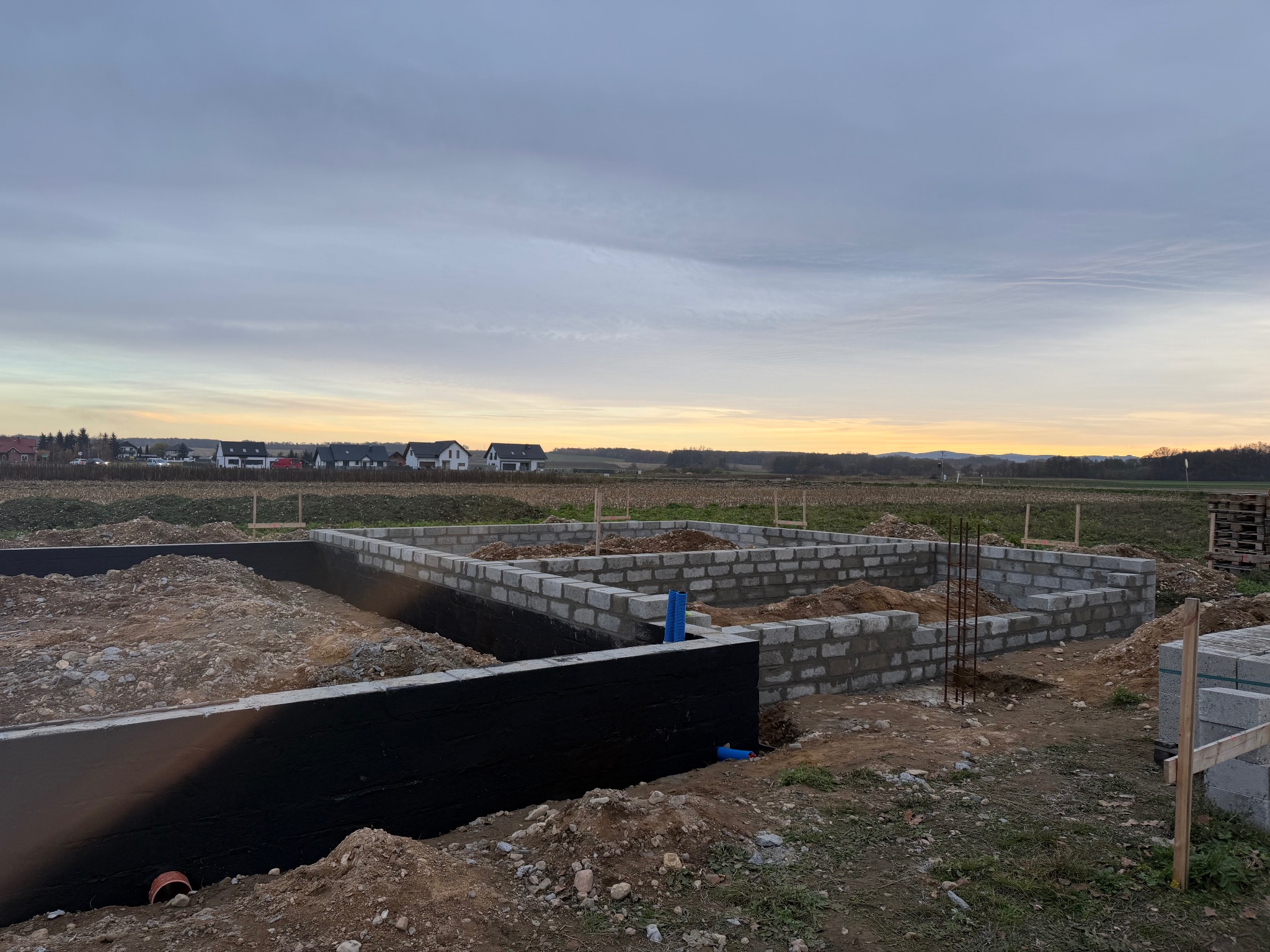 Fundamenty domu w trakcie budowy: ściany z bloczków betonowych, izolacja przeciwwilgociowa, zbrojenie. W tle krajobraz wiejski i zachodzące słońce.