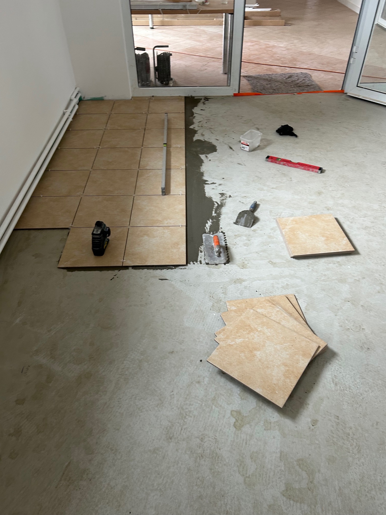 Układanie beżowych płytek podłogowych w pomieszczeniu, widoczne narzędzia glazurnicze i poziomica, częściowo ułożona podłoga, etap prac wykończeniowych.