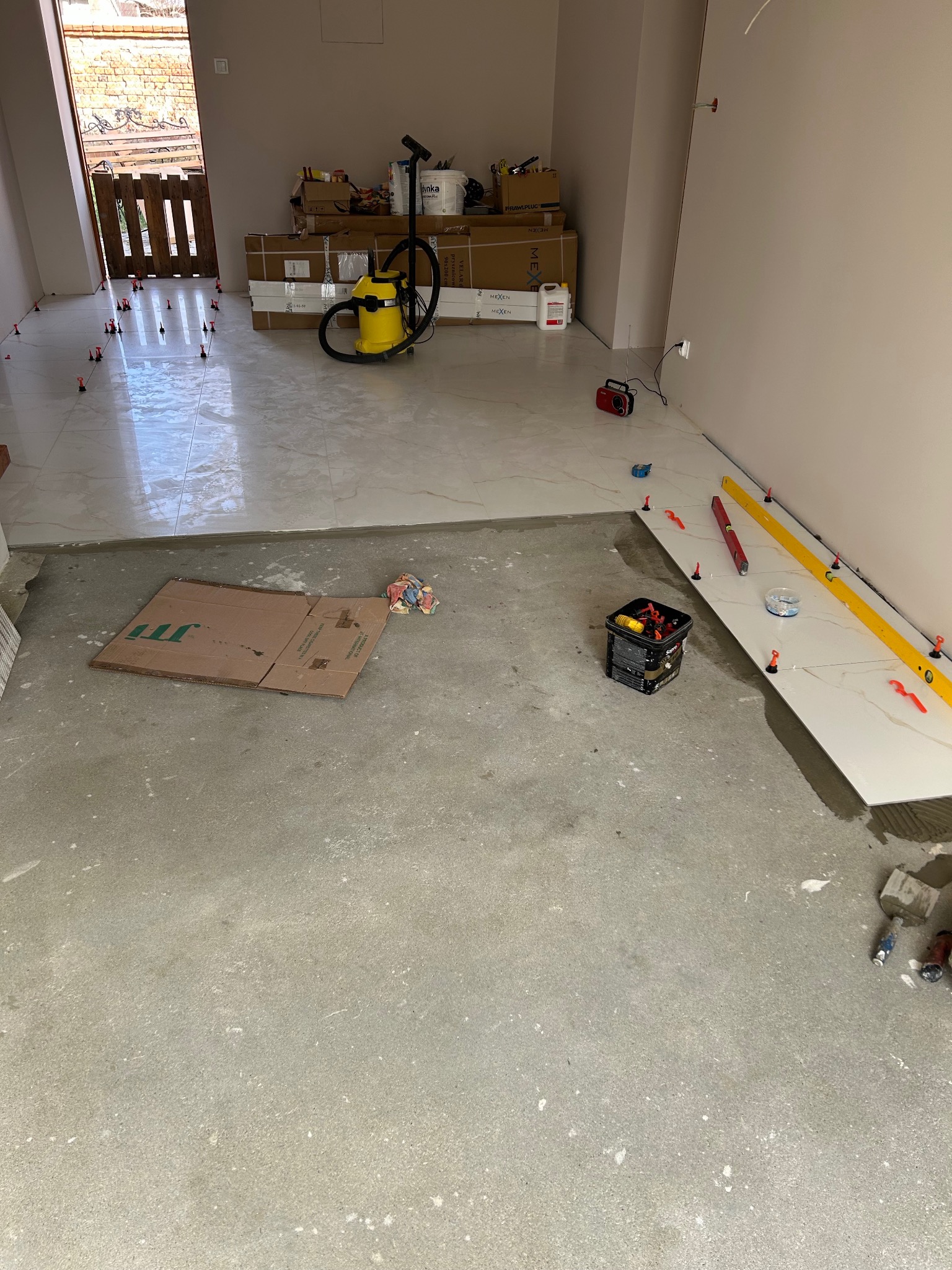 Układanie płytek podłogowych w jasnym wnętrzu. Widoczne poziomice, krzyżyki dystansowe i narzędzia glazurnicze. Częściowo ułożona podłoga z płytek imitujących marmur.