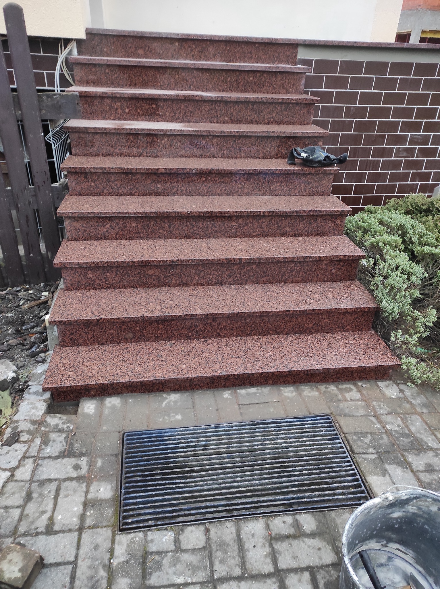 Granitowe schody zewnętrzne z brązowego kamienia, prowadzące do budynku z ceglaną elewacją. Na schodach leżą robocze rękawice, a obok rośnie krzew.
