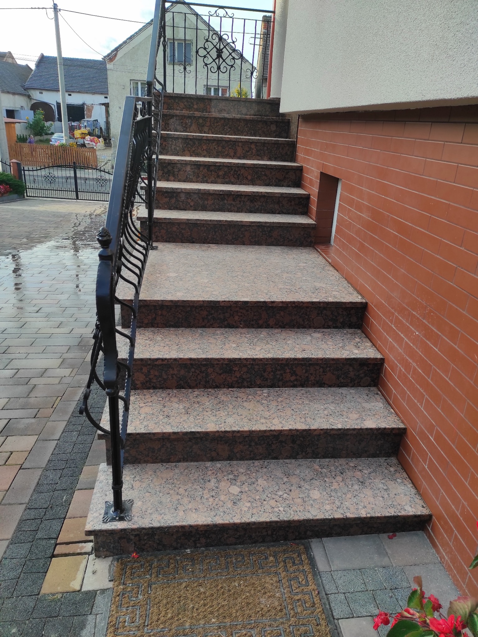 Granitowe schody zewnętrzne z czarną, kutą balustradą, prowadzące do budynku z ceglaną elewacją. Widoczny fragment chodnika z kostki brukowej i wycieraczka.