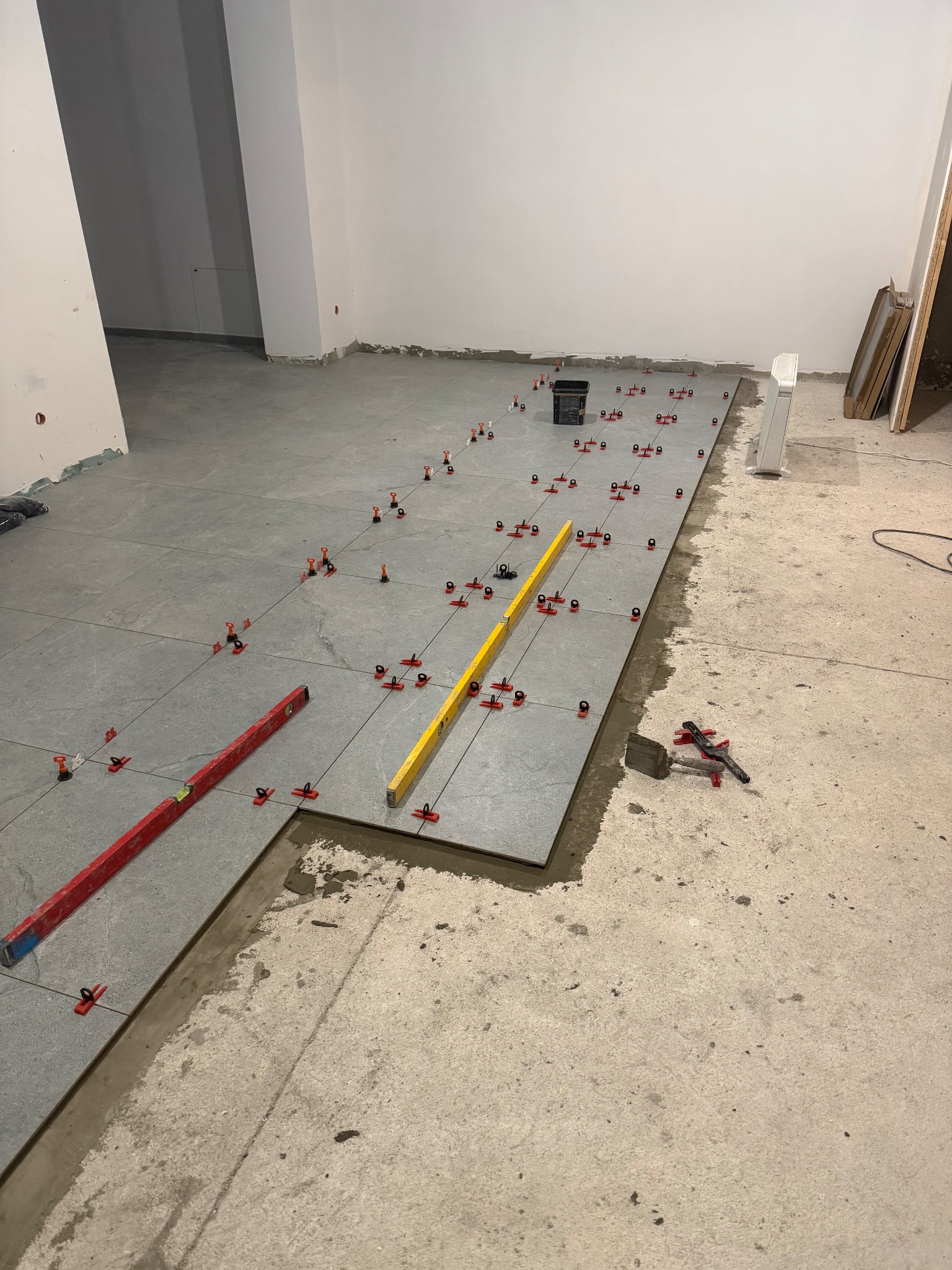 Układanie szarych płytek podłogowych z użyciem poziomnic i klipsów poziomujących. Widoczne narzędzia glazurnicze i fragment surowej posadzki.