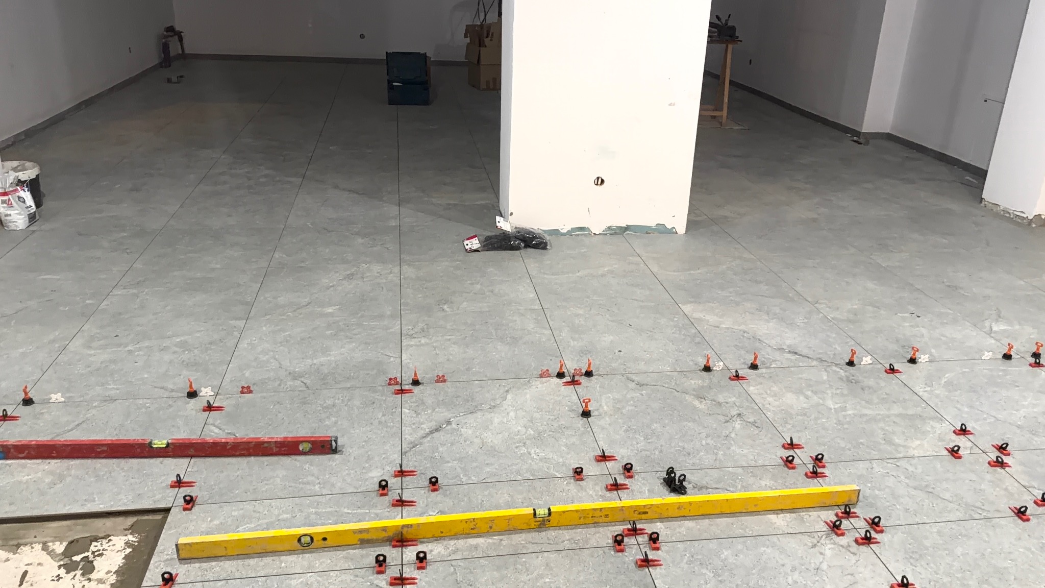 Szare płytki podłogowe w trakcie układania, z użyciem poziomnic i systemu poziomowania. Widoczny fragment nieukończonej podłogi i biały filar w tle.