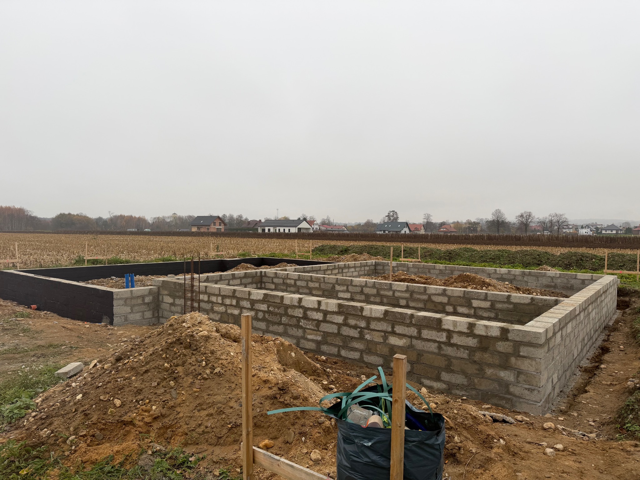 Fundamenty domu z bloczków betonowych na działce, widoczne zbrojenia i izolacja. W tle zabudowa jednorodzinna i pochmurne niebo.