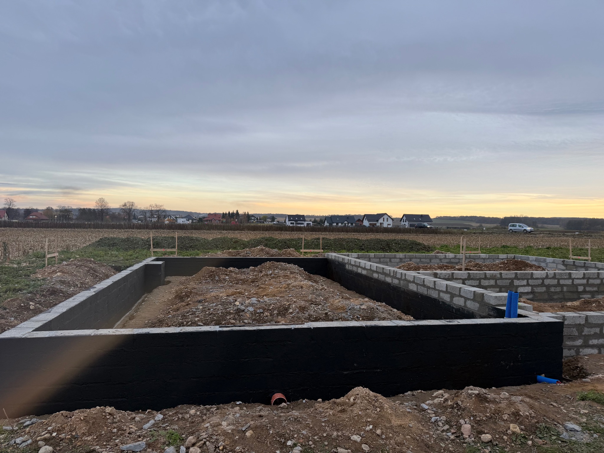 Fundamenty domu w trakcie budowy: czarna izolacja pionowa, częściowo wymurowane ściany z bloczków, ziemia wewnątrz, krajobraz w tle, pochmurne niebo.