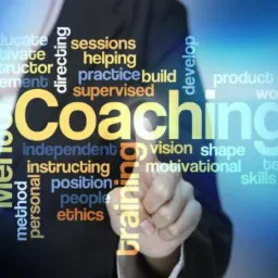 Dłoń wskazująca na słowo Coaching otoczone innymi słowami związanymi z rozwojem osobistym i edukacją, na niebieskim tle.